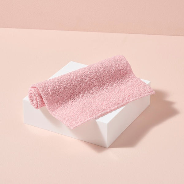 Rolled pink exfoliating towel on a white block against a beige background