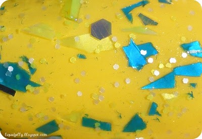 A close-up view of multicolored glitter nail polish swatch with turquoise and yellow shards mixed with small yellow hex glitter painted over yellow nail polish.
